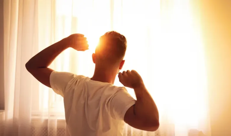 Homem espreguiçando-se pela manhã diante de uma janela iluminada pelo sol.