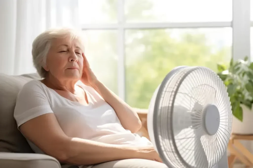 Idosa sentada em sofá, sentindo desconforto devido ao calor, com ventilador próximo