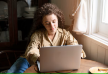 Mulher com cabelo cacheado usando um notebook em uma mesa de madeira perto da janela.