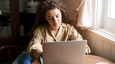 Mulher com cabelo cacheado usando um notebook em uma mesa de madeira perto da janela.