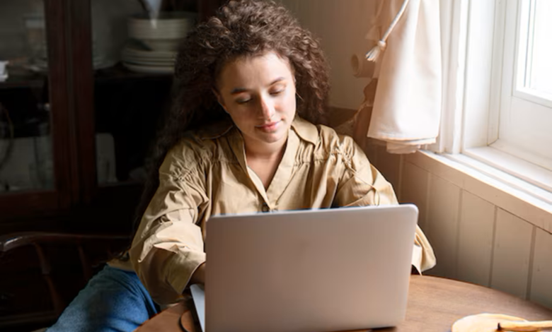 Mulher com cabelo cacheado usando um notebook em uma mesa de madeira perto da janela.