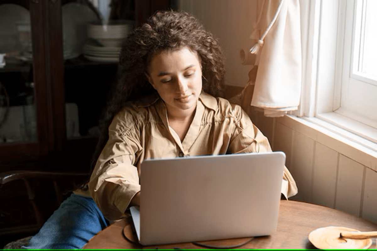 Mulher com cabelo cacheado usando um notebook em uma mesa de madeira perto da janela.