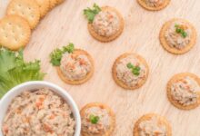 Biscoitos crocantes com patê de frango cremoso, decorados com cenoura e cebolinha, servidos sobre uma tábua de madeira.