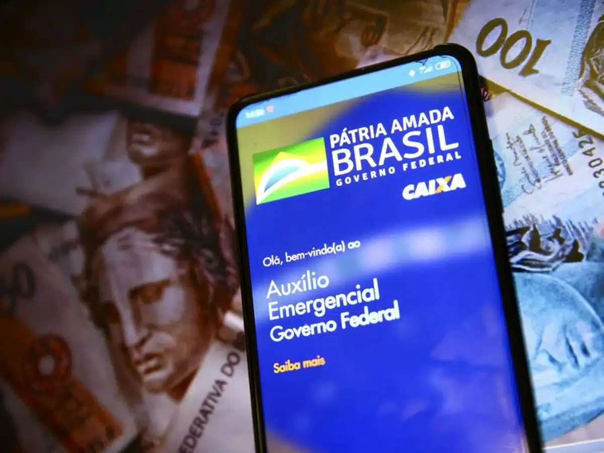Tela de celular mostrando o aplicativo do Auxílio Emergencial com fundo de cédulas de dinheiro