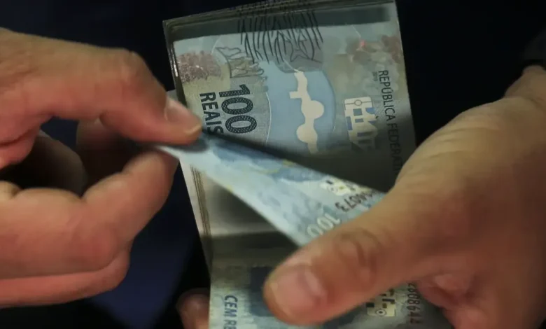 Mãos contando cédulas de 100 reais, representando pagamento de benefícios.