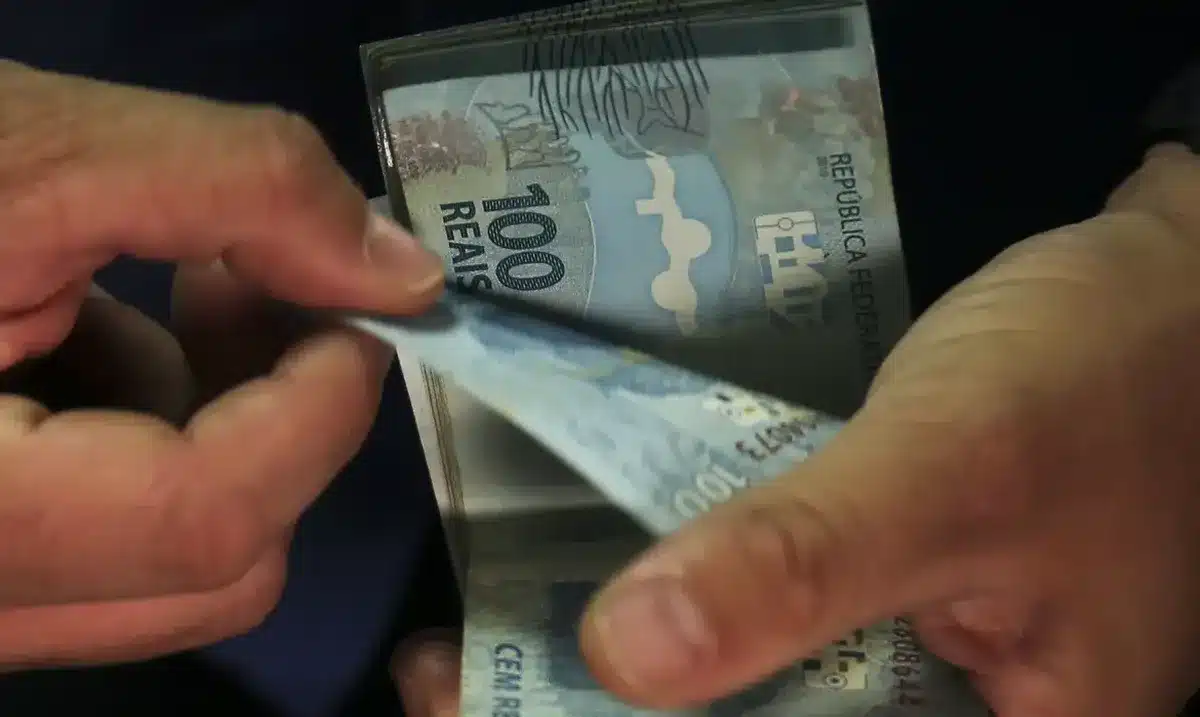 Mãos contando cédulas de 100 reais, representando pagamento de benefícios.