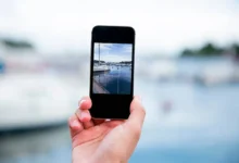 Mão segurando um smartphone tirando uma foto de um cenário ao ar livre com barcos e água ao fundo