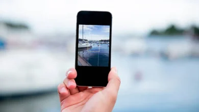 Mão segurando um smartphone tirando uma foto de um cenário ao ar livre com barcos e água ao fundo