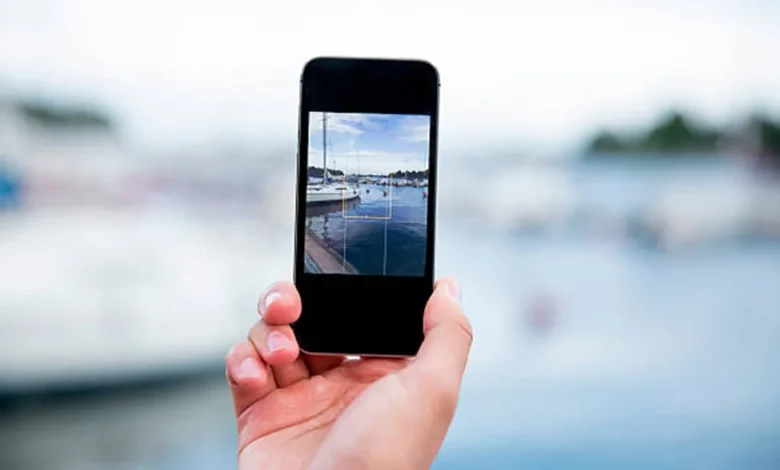 Mão segurando um smartphone tirando uma foto de um cenário ao ar livre com barcos e água ao fundo