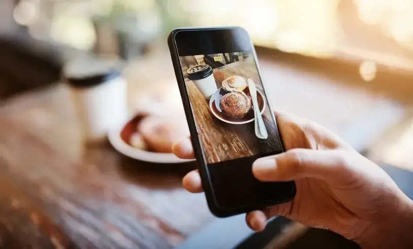 Pessoa segurando um smartphone e fotografando um café da manhã em uma mesa de madeira