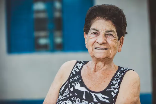  Mulher idosa sorrindo ao ar livre, vestindo roupa estampada.