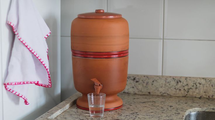 Filtro de barro com detalhes vermelhos, colocado sobre uma bancada de granito, com um copo de água ao lado.