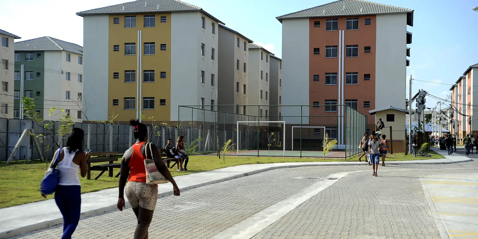 "Pessoas caminhando em uma área residencial com prédios e campo de futebol, ilustrando a oportunidade de conquistar a casa própria."