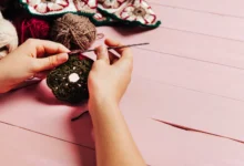 Mãos segurando um trabalho de crochê em cores verde e vermelha com fundo de madeira rosa