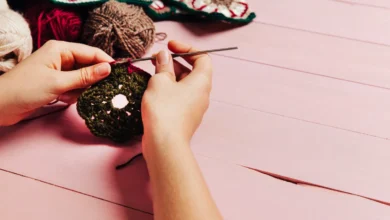 Mãos segurando um trabalho de crochê em cores verde e vermelha com fundo de madeira rosa