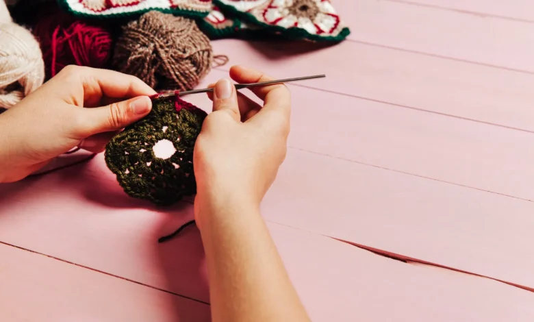 Mãos segurando um trabalho de crochê em cores verde e vermelha com fundo de madeira rosa