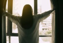 Mulher de costas abrindo as cortinas de uma janela com vista para a cidade, simbolizando gratidão e novos começos