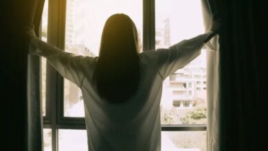 Mulher de costas abrindo as cortinas de uma janela com vista para a cidade, simbolizando gratidão e novos começos
