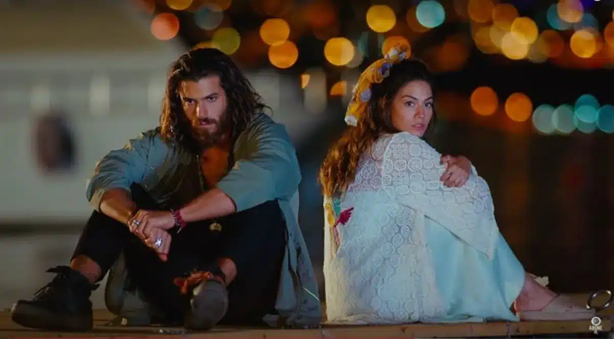 Casal de protagonistas de Erkenci Kuş sentados em um píer, com luzes ao fundo