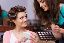 Mulher sorrindo enquanto vê seu reflexo em um celular, com cabelo preso em bigudi e outra mulher preparando maquiagem para o Dia das Mães