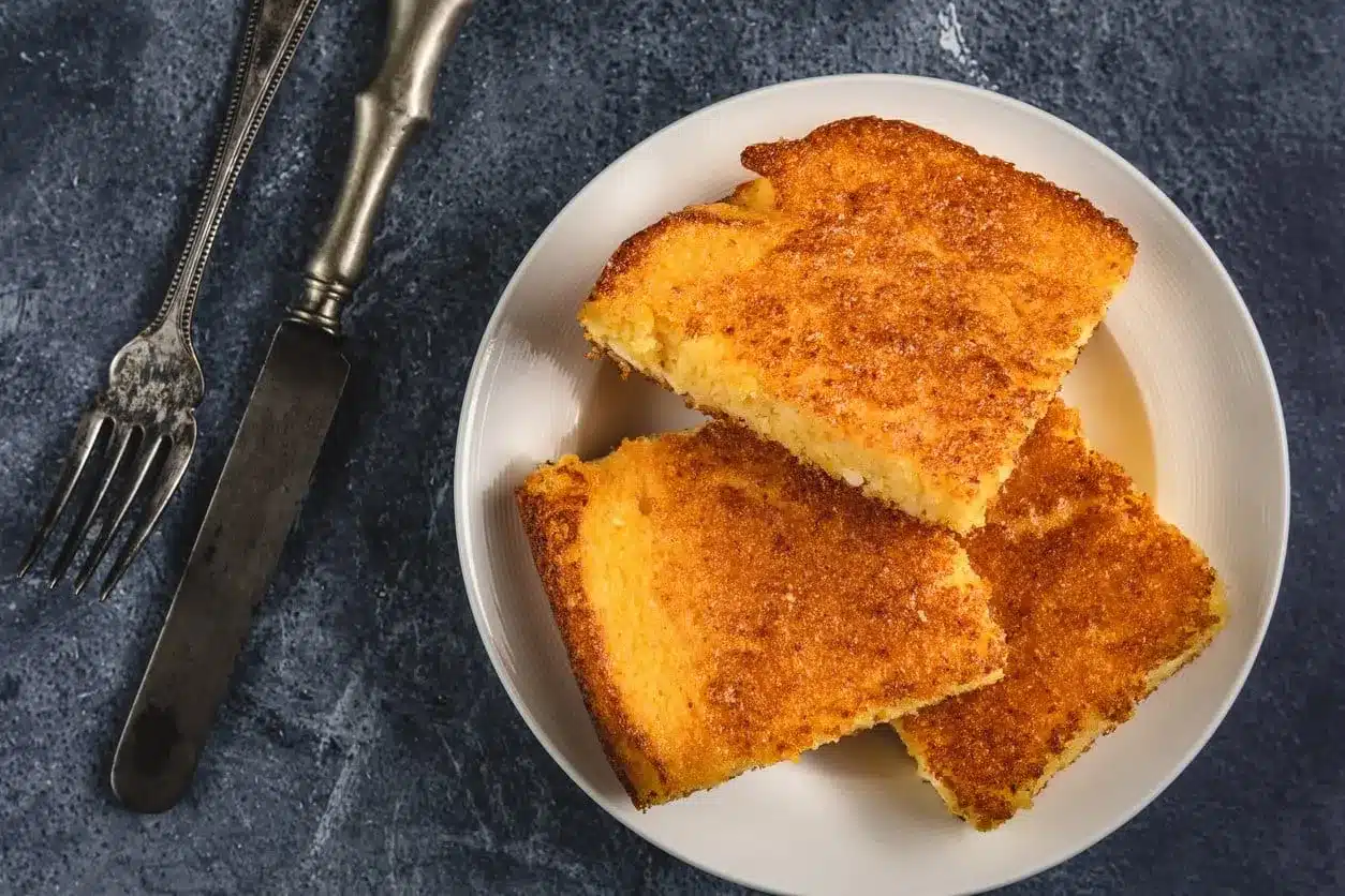 Imagem de fatias de pão de cuscuz douradas, servidas com talheres, prontas para o café da manhã ou lanche da tarde