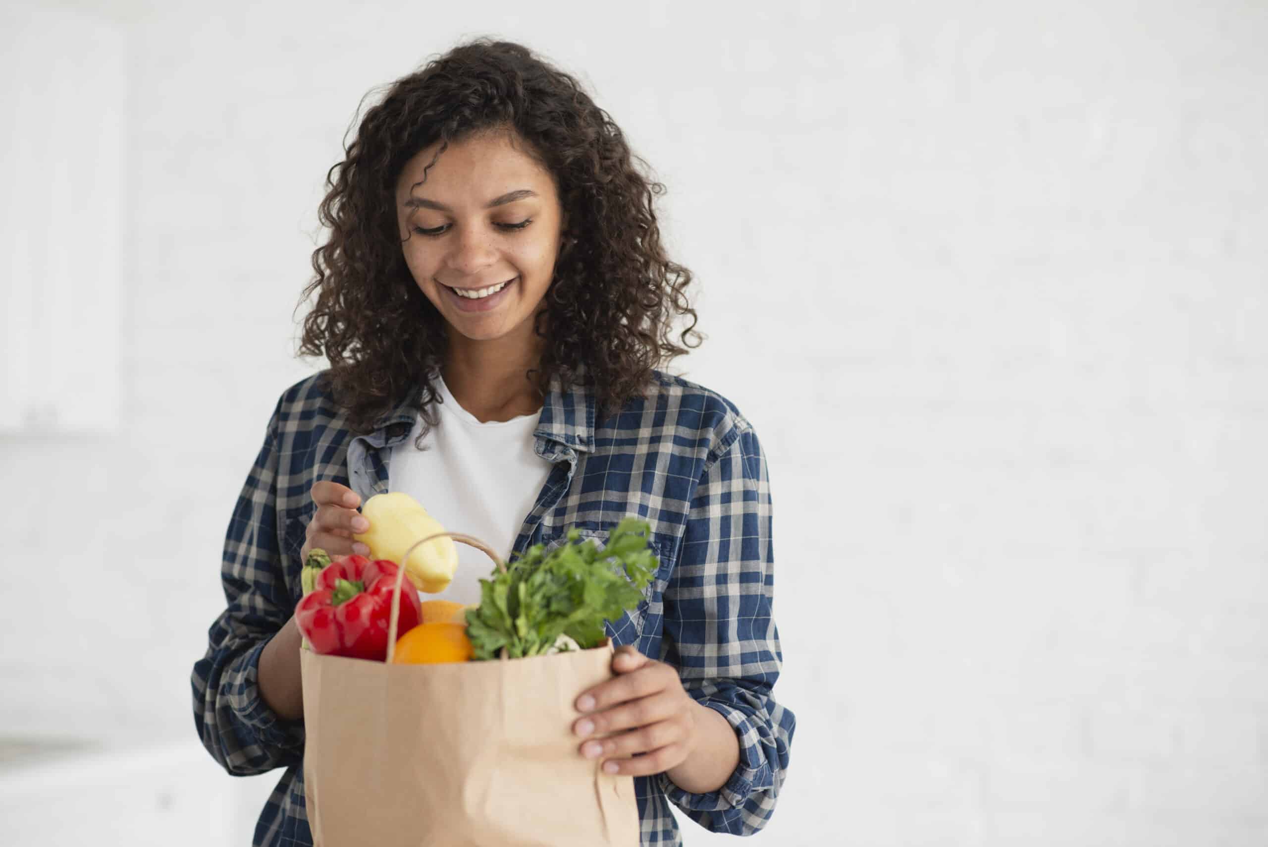 Mulher sorrindo enquanto observa frutas e vegetais frescos em uma sacola de papel.