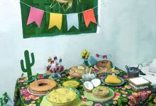 Mesa decorada para festa junina com toalha floral, comidas típicas e bandeirolas coloridas.