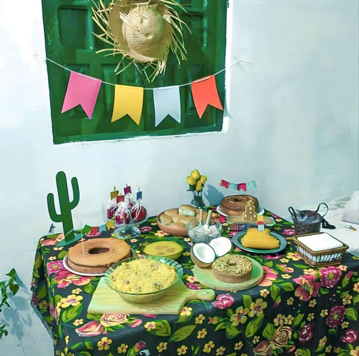 Mesa decorada para festa junina com toalha floral, comidas típicas e bandeirolas coloridas.