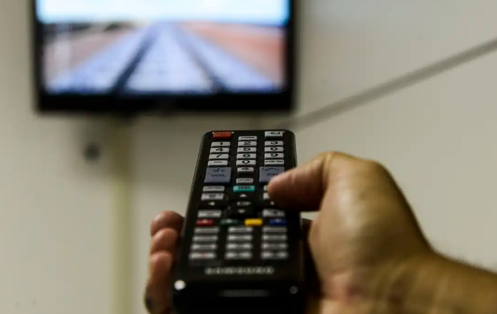 Pessoa usando controle remoto diante de televisão com sinal de TV digital ao fundo.