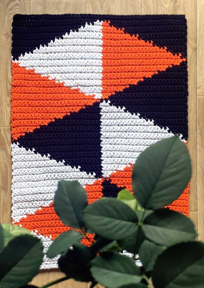 Tapete de crochê com formas geométricas em laranja, branco e preto, sobre piso de madeira, com folhas verdes em primeiro plano.