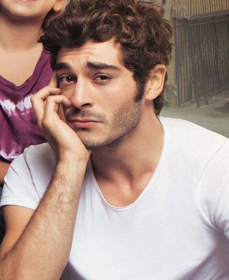 Ator turco Burak Deniz com expressão pensativa, usando camiseta branca, em ambiente interno.