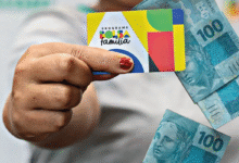 Cartão do programa Bolsa Família sendo segurado por uma mão, acompanhado por notas de 100 reais.