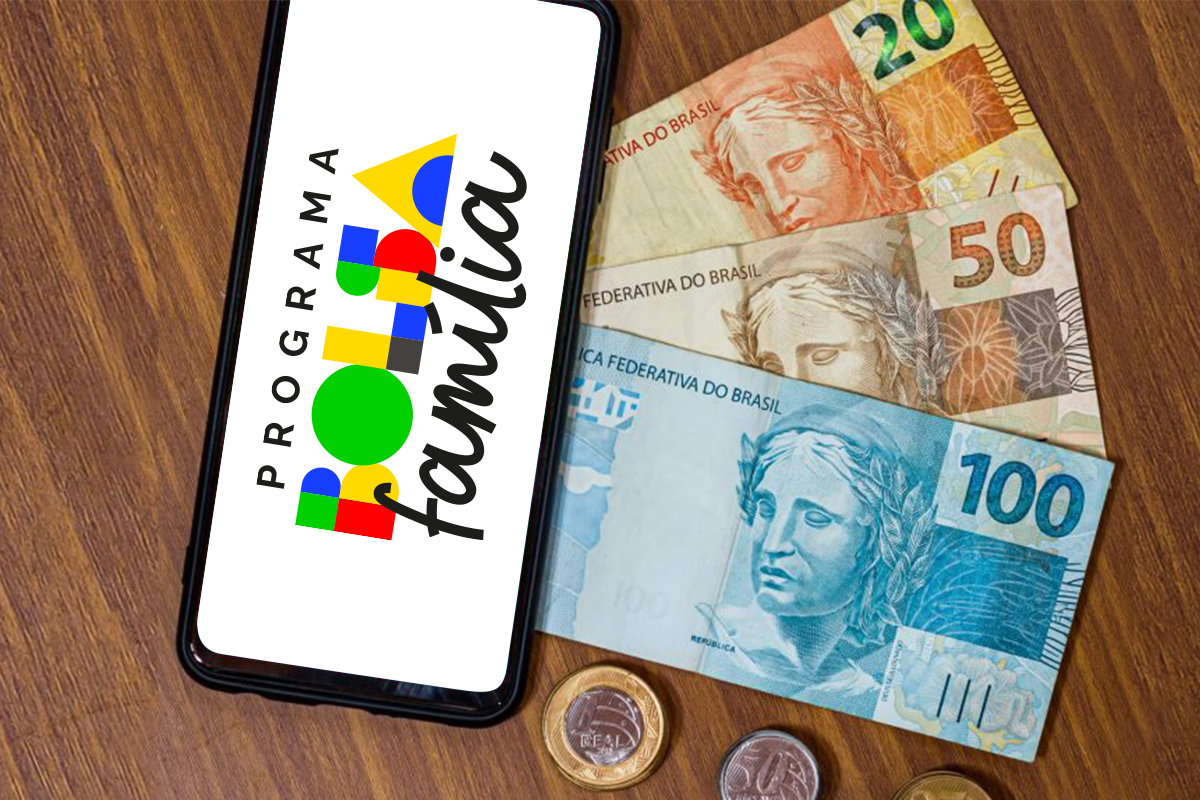 Celular mostrando o logo do Programa Bolsa Família ao lado de cédulas e moedas do Real brasileiro sobre uma mesa de madeira
