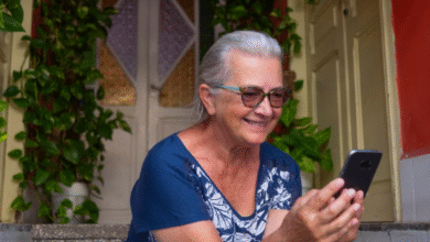 Mulher sorridente usando o celular sentada em frente à sua casa.