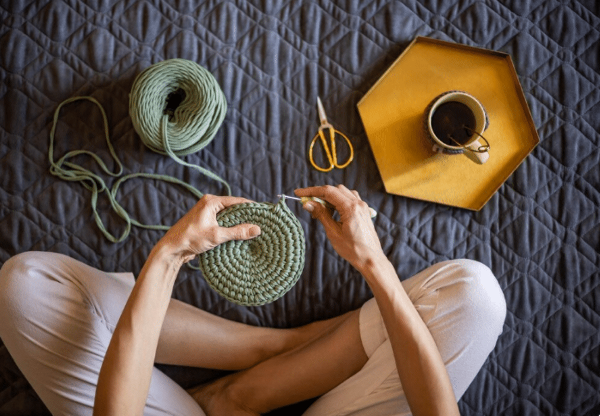 Pessoa fazendo crochê sentada, com material de trabalho e uma xícara de chá ao lado.