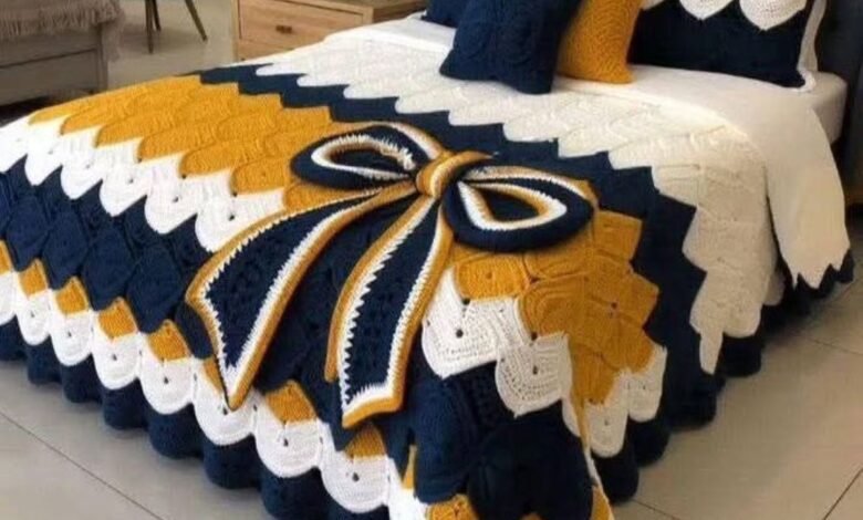 Jogo de cama em crochê nas cores azul, mostarda e branco com laço decorativo sobre a cama.