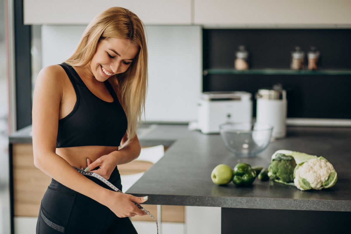 Mulher sorrindo e medindo a cintura enquanto se alimenta de alimentos saudáveis, como brócolis e maçã, em sua cozinha
