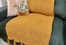 Manta de crochê amarela com franjas sobre sofá verde e almofada decorativa com estampa moderna.