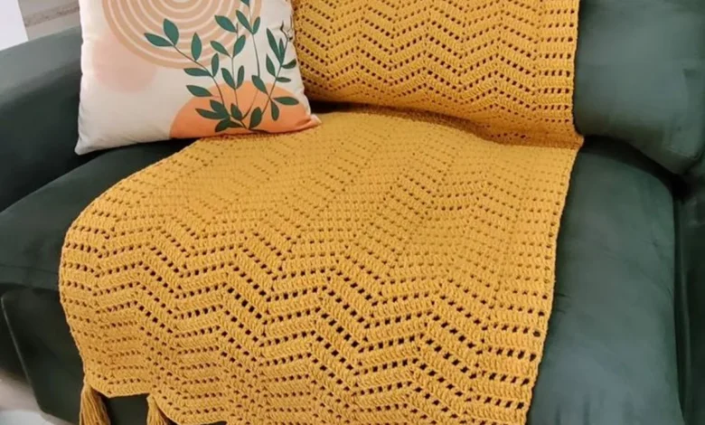 Manta de crochê amarela com franjas sobre sofá verde e almofada decorativa com estampa moderna.