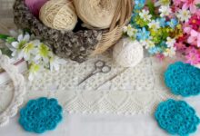 Peças de crochê em tons neutros e azul turquesa, com novelos de lã e flores coloridas ao redor, destacando artesanato manual.