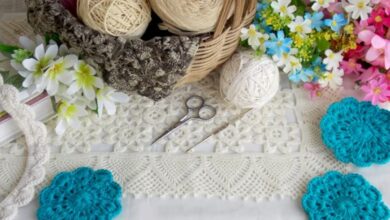Peças de crochê em tons neutros e azul turquesa, com novelos de lã e flores coloridas ao redor, destacando artesanato manual.