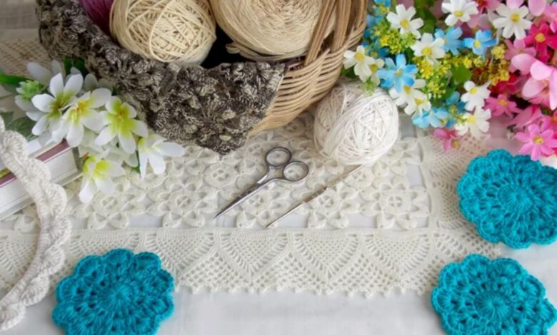 Peças de crochê em tons neutros e azul turquesa, com novelos de lã e flores coloridas ao redor, destacando artesanato manual.