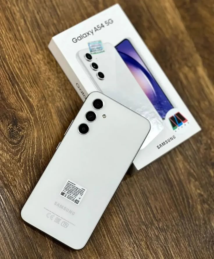 Smartphone Samsung Galaxy A54 branco com câmera tripla, visto de costas sobre uma mesa de madeira com a caixa ao fundo.