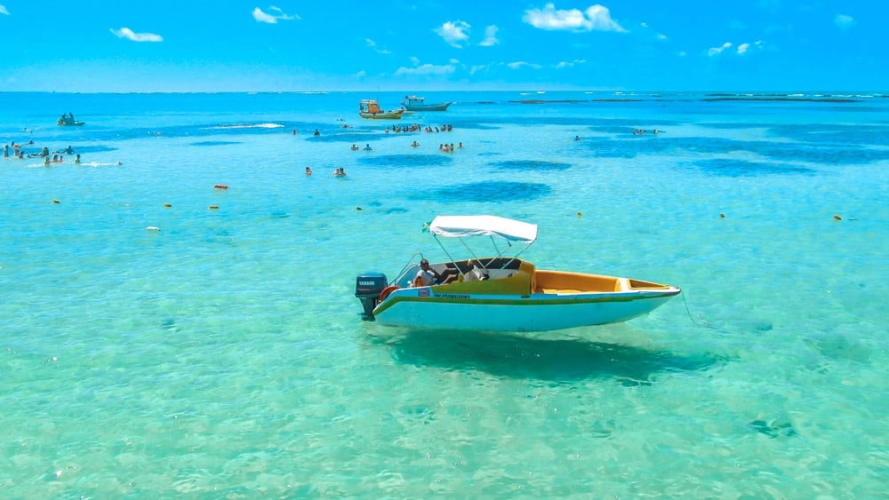 Barco flutuando sobre as águas cristalinas da Costa dos Corais em Maceió, com pessoas aproveitando o mar ao fundo