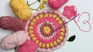 Detalhe de peça redonda em crochê colorido com fios rosa, amarelo e marrom ao redor, pronta para acabamento.