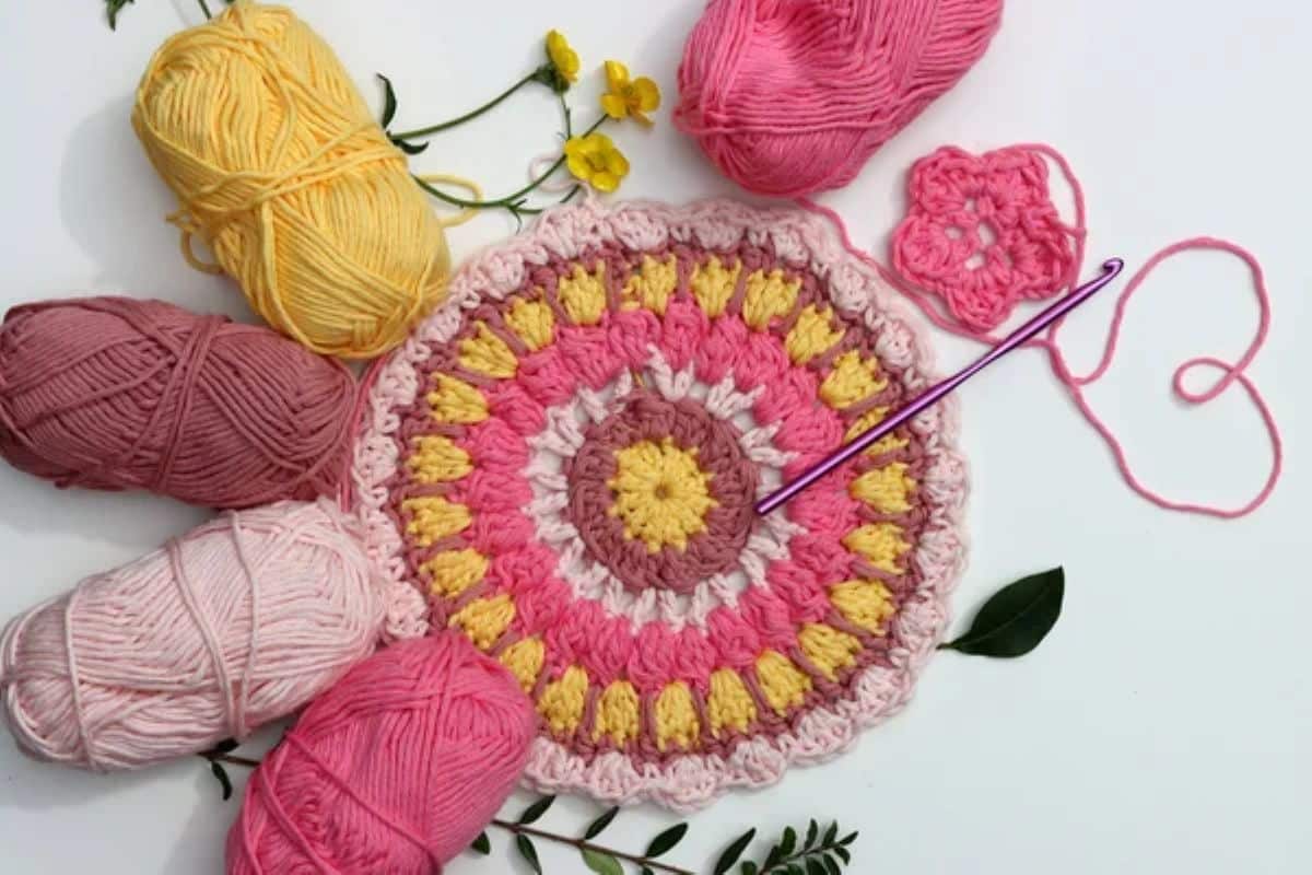 Detalhe de peça redonda em crochê colorido com fios rosa, amarelo e marrom ao redor, pronta para acabamento.