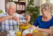 Casal de idosos sorrindo enquanto prepara uma salada saudável na mesa do café da manhã.