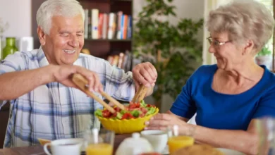 Casal de idosos sorrindo enquanto prepara uma salada saudável na mesa do café da manhã.