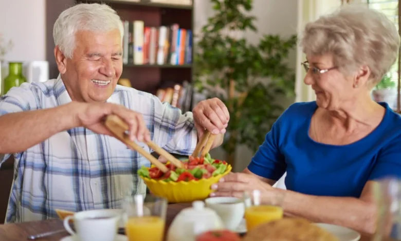 Casal de idosos sorrindo enquanto prepara uma salada saudável na mesa do café da manhã.
