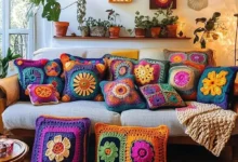 Almofadas de crochê coloridas e detalhadas sobre sofá cinza em sala iluminada, com plantas ao fundo.